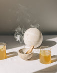 Kleine Räucherschale aus beigefarbenem Travertinstein mit Palo Santo Stick und Globe Räucherkugel aus Travertin mit Kruut Oxymel Drinks