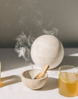 Kleine Räucherschale aus beigefarbenem Travertinstein mit Palo Santo Stick und Globe Räucherkugel aus Travertin mit Kruut Oxymel Drinks