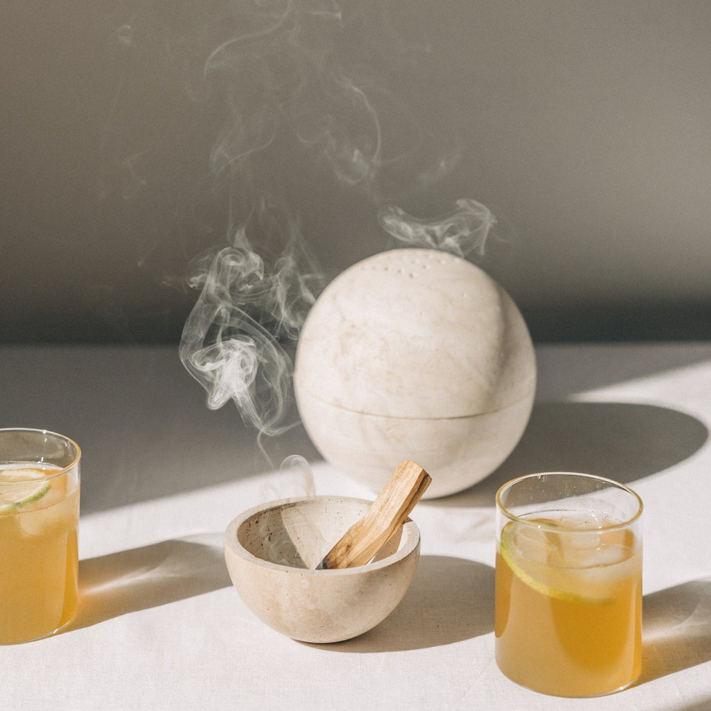 Kleine Räucherschale aus beigefarbenem Travertinstein mit Palo Santo Stick und Globe Räucherkugel aus Travertin mit Kruut Oxymel Drinks